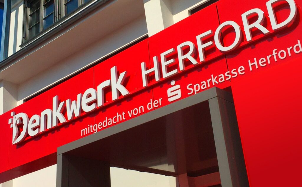 Das moderne Design des Denkwerk Herford zeigt sich im Eingangsbereich mit einem hellen roten Vordach und dem Firmennamen, mitgetragen von der Sparkasse Herford.