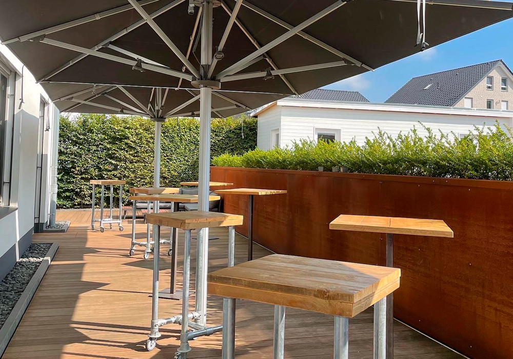 Eine sonnengeschützte Terrasse mit stylischen Holztischen und Barhockern, die zum Networking in der entspannten Atmosphäre des Denkwerk Herford einlädt.