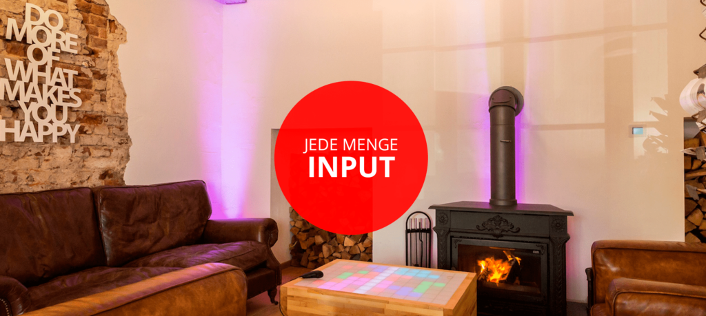 Bild eines gemütlichen Raums mit einer Ledercouch und einem Kamin, der ein Feuer entfacht. An der Wand hängt eine inspirierende Wanddekoration mit dem Schriftzug "DO MORE OF WHAT MAKES YOU HAPPY". Über einem Tisch mit buntem Lichtkreis im Vordergrund steht der Text "JEDE MENGE INPUT".