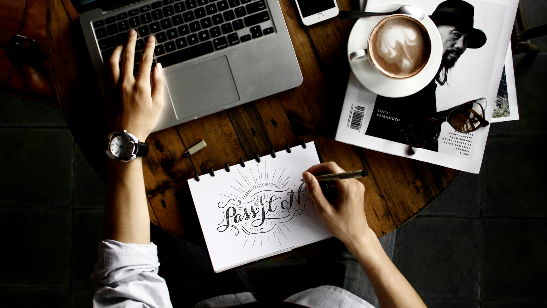 Person gestaltet kreatives Lettering in einem Notizbuch mit dem Slogan 'Pass it on', neben einem Laptop und Design-Inspirationen, im Kontext der Findung eines Unternehmensnamens.