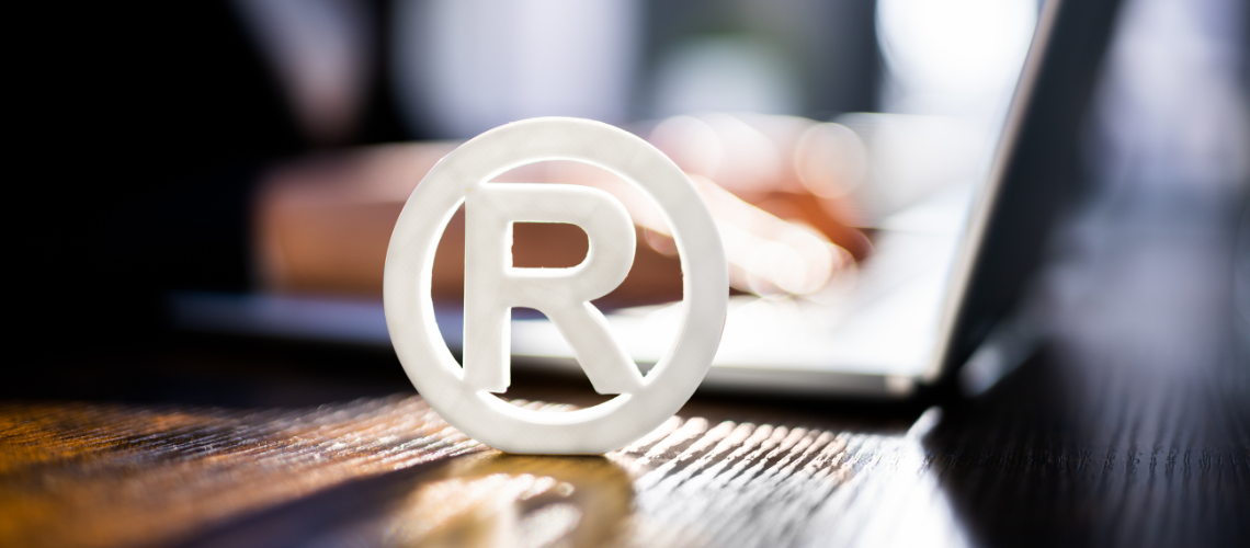 Das "R" für das eingetragene Markenzeichen im Vordergrund, mit unscharfem Hintergrund, der eine Person bei der Arbeit an einem Laptop zeigt, was auf Tätigkeiten bezogen auf das Handelsregister hindeuten könnte.