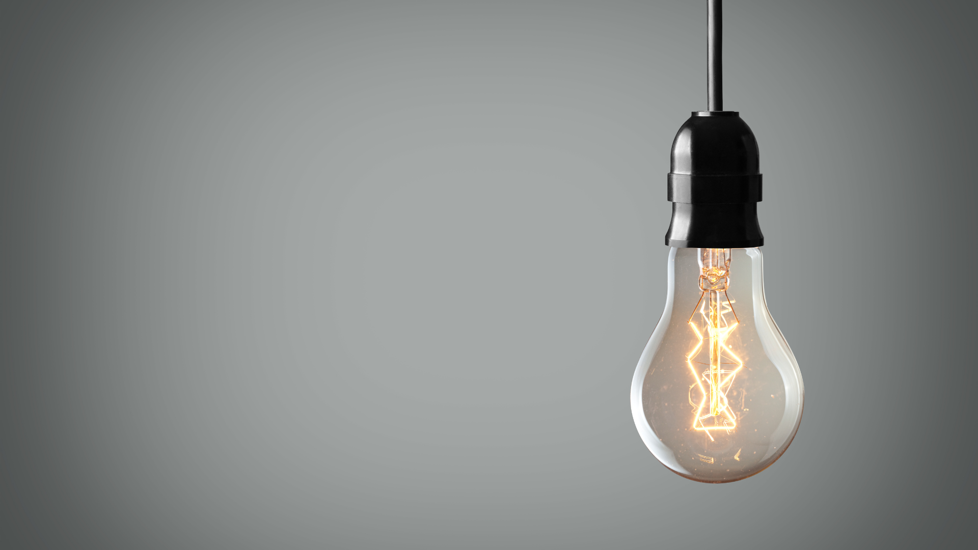 Glühbirne mit leuchtendem Filament hängt vor einem grauen Hintergrund, Symbol für Ideenfindung und Inspiration, im Zusammenhang mit 'Nebenberufliche Tätigkeit: ein Leitfaden'