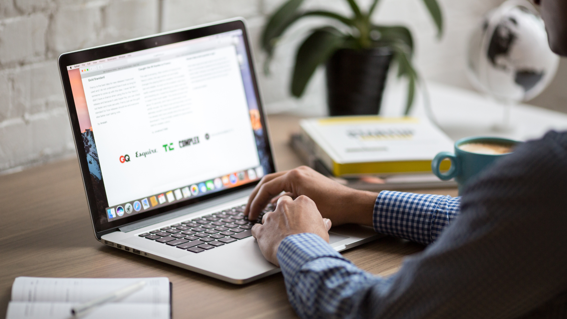 Person in einem karierten Hemd tippt auf einem Laptop, auf dem Bildschirm sind Text und Logos zu sehen, neben einer Kaffeetasse, Pflanze und Büchern, die sich auf 'Die perfekte Domain auswählen: ein Leitfaden' beziehen.