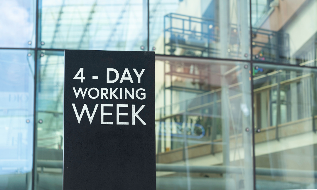 Ein Schild vor einem modernen Bürogebäude verkündet '4-Day Working Week' mit einem klaren Hintergrund aus Glasfassaden und Innenstrukturen.