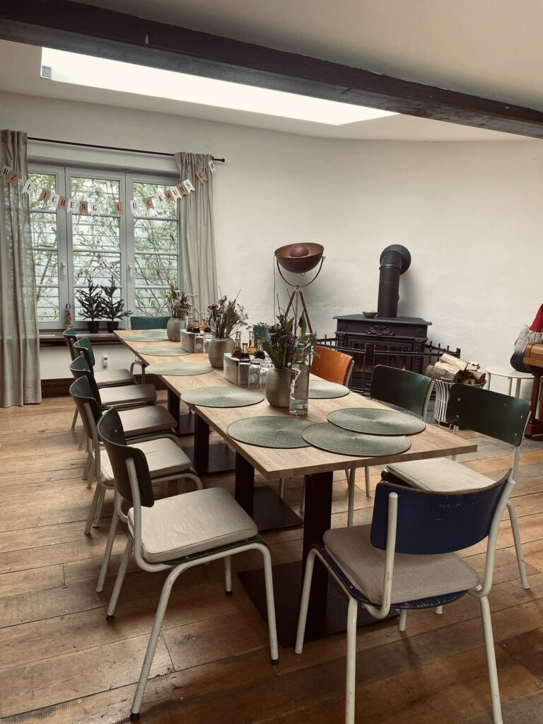 Ein stilvoll eingerichteter Besprechungs- oder Veranstaltungsraum in der Alten Sackfabrik mit rustikalem Holzboden, einem langen Holztisch, Vintage-Stühlen, Blumenarrangements und einem Kaminofen im Hintergrund.