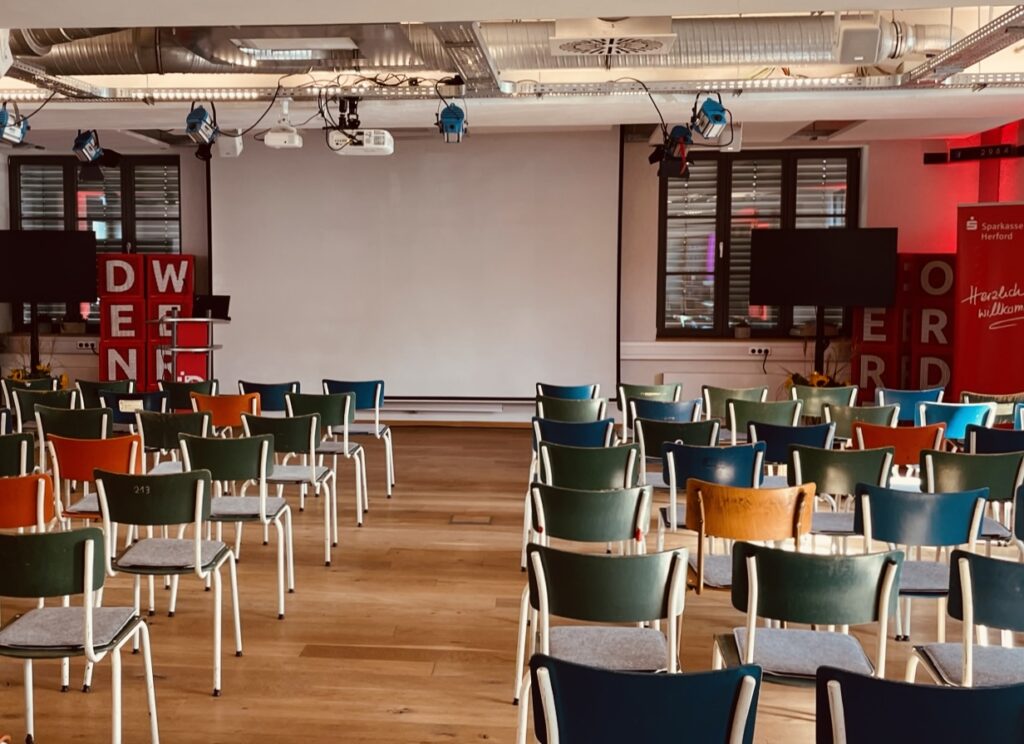 Keynote Veranstaltungsraum mit großer Leinwand, moderner Beleuchtung, farbigen Stühlen und Technik für Vorträge, Seminare und Präsentationen