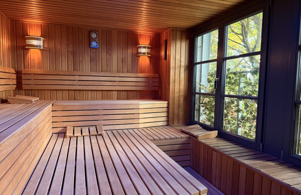 Innenansicht einer modernen Sauna aus hellem Holz mit großen Fenstern, Sitzbänken und stimmungsvoller Beleuchtung