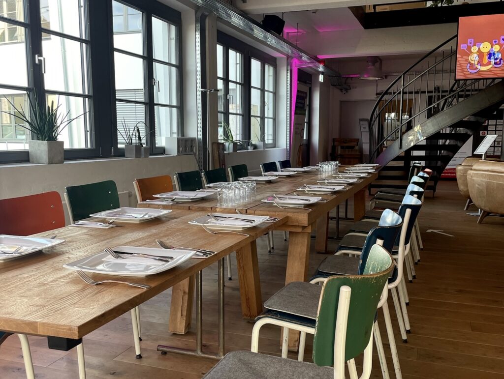 Lange gedeckte Holztische mit bunten Stühlen in der hellen Lobby des Denkwerks Herford mit großen Fenstern und Industrielampen