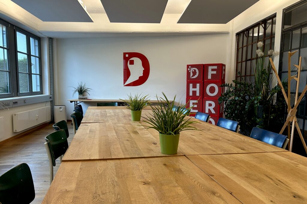 Besprechungsraum Brainforce Three im Denkwerk Herford mit großem Holztisch, grünen Pflanzen, Fensterfront und modernem Design – ideal für Workshops und Seminare.