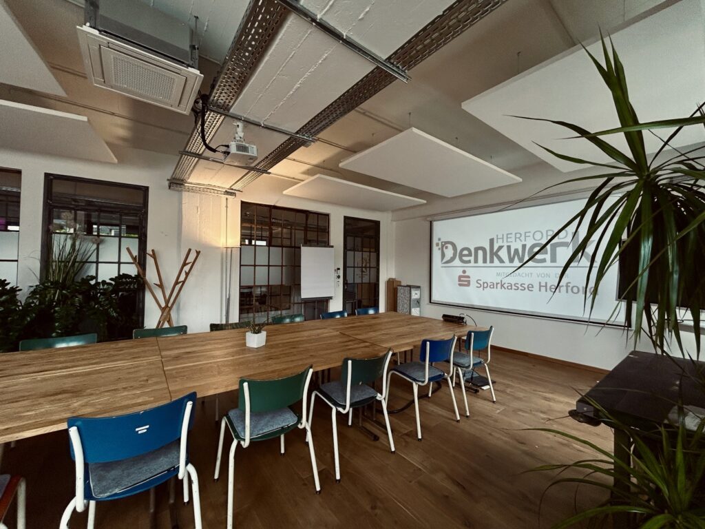 Großer Seminar- und Konferenzraum im Denkwerk Herford mit flexibel angeordneten Konferenztischen, Leinwand, Beamer und moderner Präsentationstechnik für Workshops, Schulungen und Tagungen.