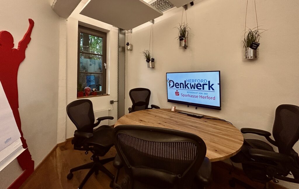 Kleiner Besprechungsraum im Denkwerk Herford mit rundem Konferenztisch, ergonomischen Bürostühlen und großem Display für Videokonferenzen, Meetings und kreative Teamarbeit.