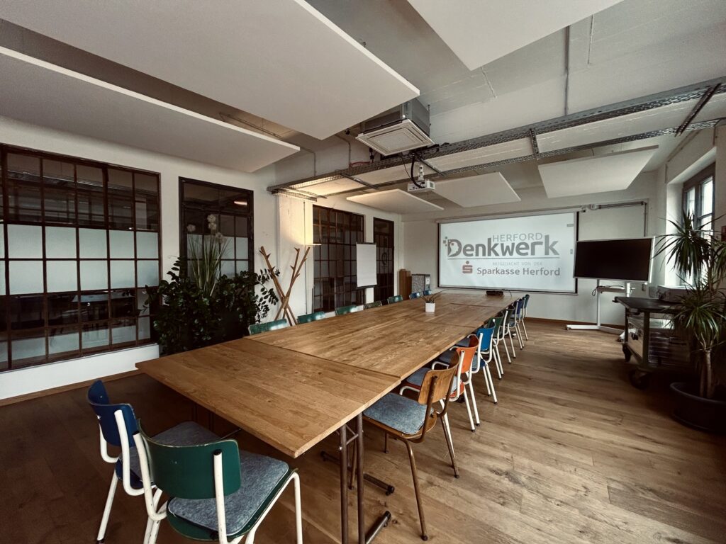 Moderner Konferenzraum im Denkwerk Herford mit großem Holztisch, flexibler Bestuhlung, Beamer, Leinwand und Präsentationstechnik – ideal für Workshops, Seminare und Meetings.
