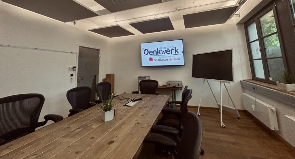 Moderner Meetingraum im Denkwerk Herford mit großem Konferenztisch, Präsentationsdisplay, interaktivem Whiteboard und ergonomischen Bürostühlen für Meetings, Workshops und Videokonferenzen.
