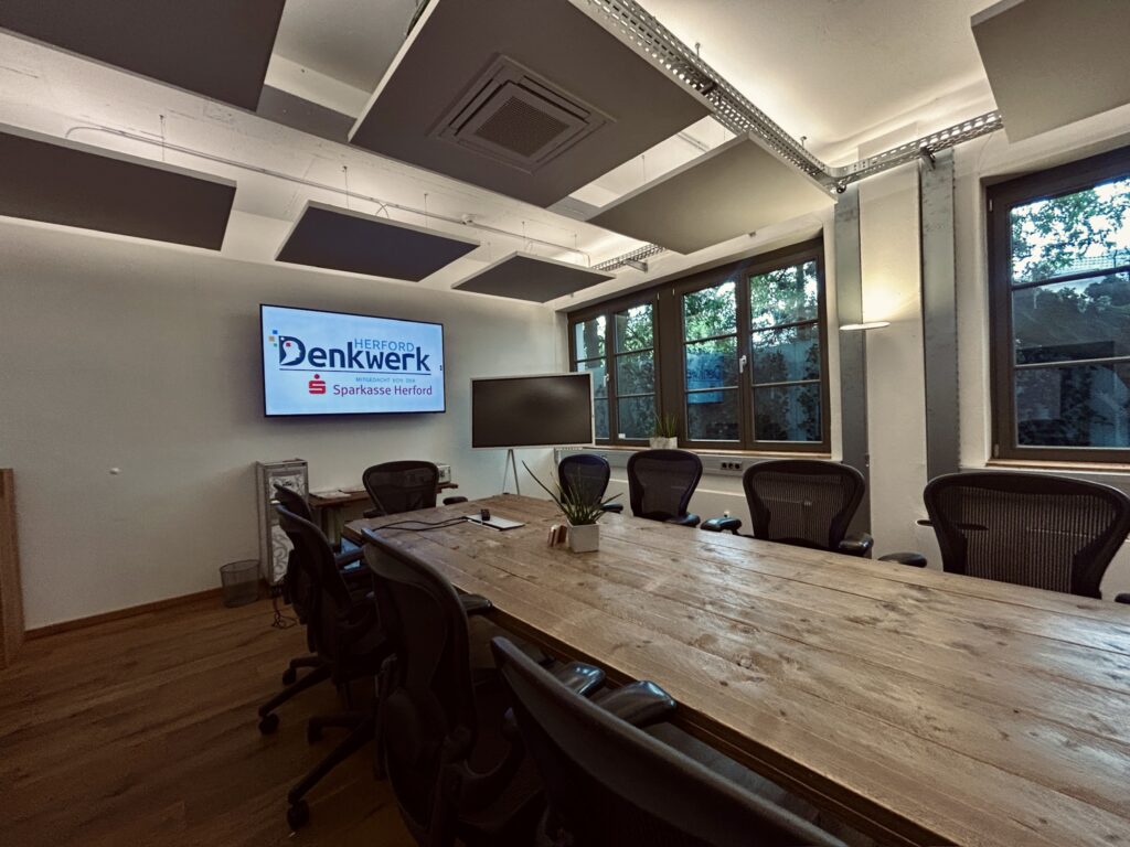 Moderner Meetingraum im Denkwerk Herford mit großem Konferenztisch, ergonomischen Bürostühlen, Präsentationsdisplay und digitalem Whiteboard für Besprechungen, Workshops und Videokonferenzen.