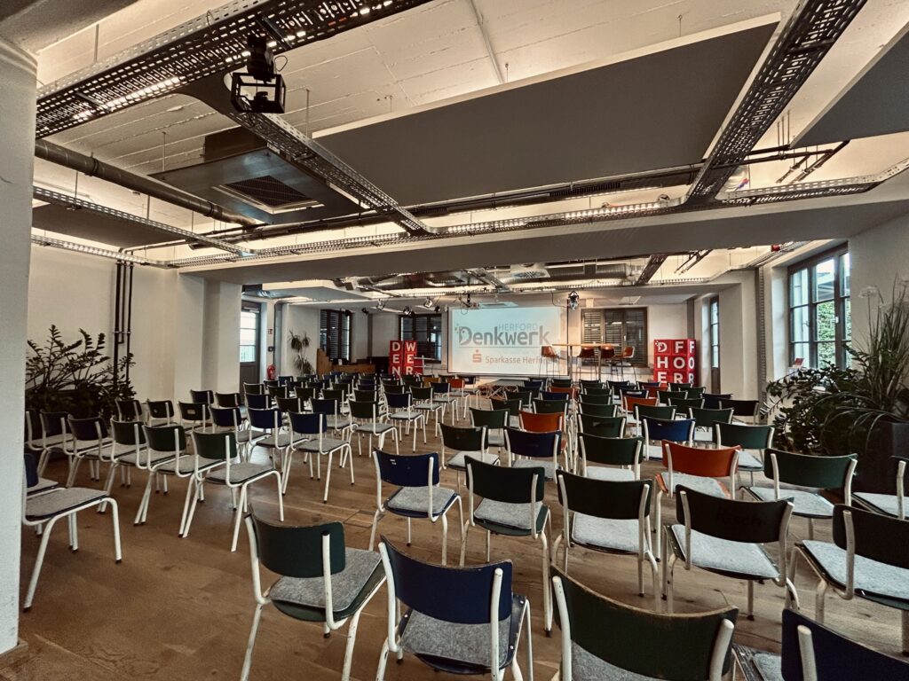 Großer Veranstaltungsraum im Denkwerk Herford mit Reihenbestuhlung, Bühne, Leinwand und moderner Präsentationstechnik – ideal für Vorträge, Tagungen, Konferenzen und Business-Events.