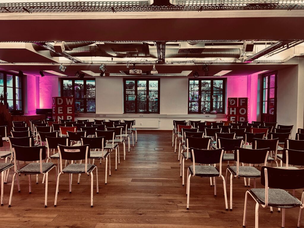 Großer Veranstaltungsraum im Denkwerk Herford mit Reihenbestuhlung für Vorträge, Präsentationen und Konferenzen. Die historische Industriearchitektur wird durch moderne Veranstaltungstechnik und stimmungsvolle LED-Beleuchtung ergänzt.